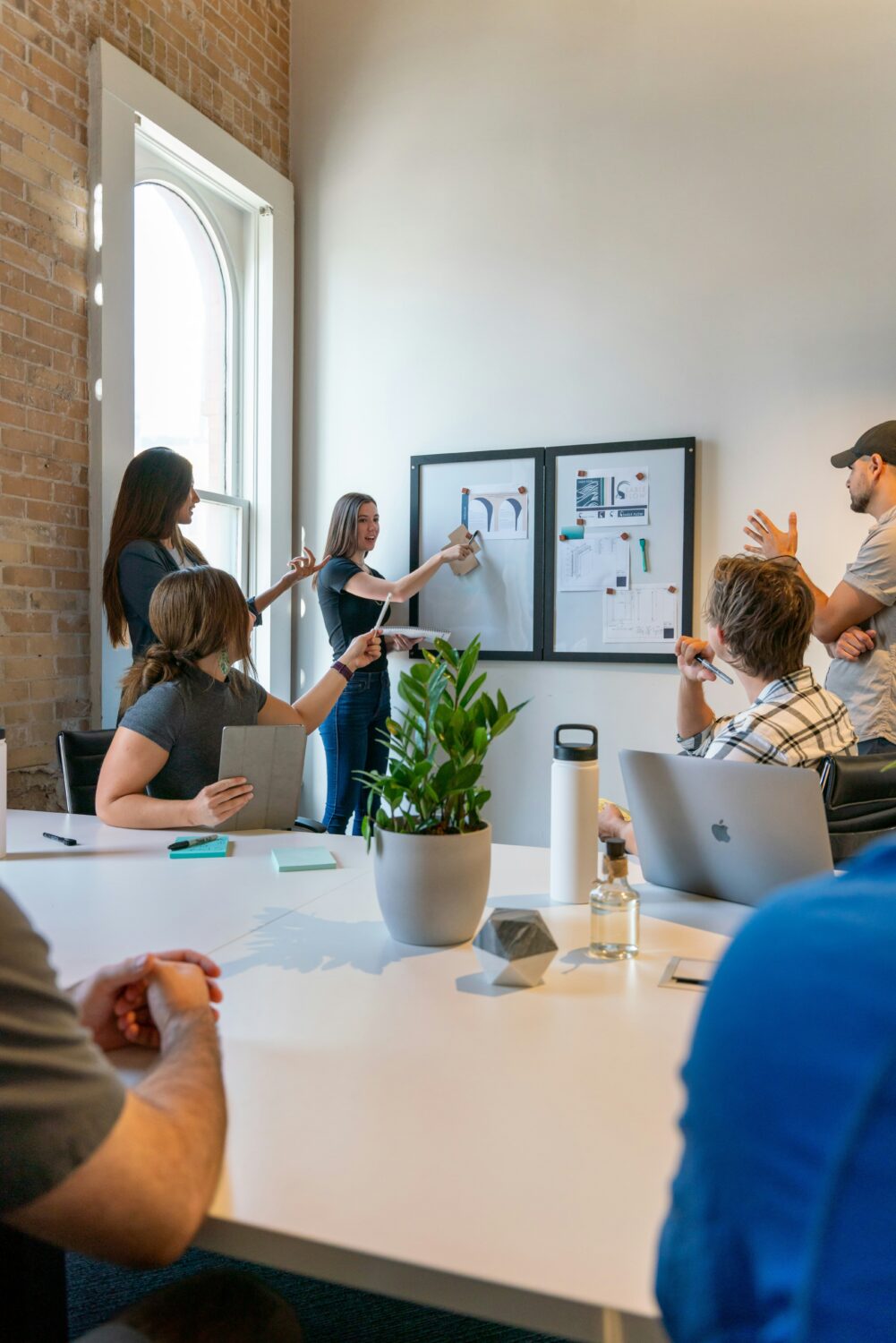 Team meets around conference table. They are in discussion as female leader stands at a whiteboard.