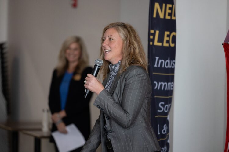 A female speaker holds a microphone and smiles while another woman smiles from the background.