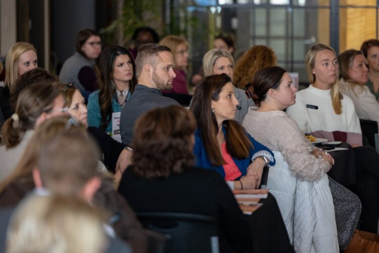 A group of professionals sits at tables watching someone at the front of a room.